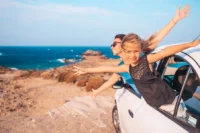 Família viajando de carro com criança na janela abrindo os braços representando dicas para viajar de carro.