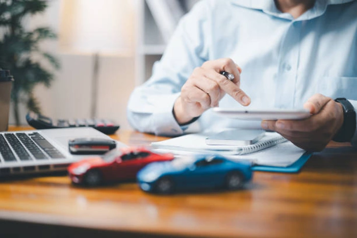 Homem faz cálculo de seguro. Sobre a mesa há dois carros em miniatura e um notebook, representando seguro cashback.