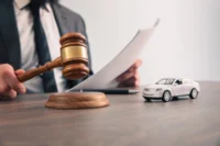 Homem simula bater com um martelo de juiz, enquanto segura documentos. Sobre sua mesa há um carro em miniatura na cor branca.