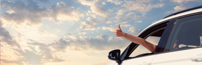 Pessoa coloca a mão para fora do veículo enquanto dirige e faz sinal de positivo. Falando sobre coberturas do seguro auto.