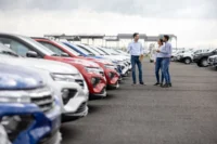 Corretor conversa com casal de compradores em meio a fileira de carros usados. Falando sobre os preço do seguro de carros usados.