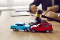 Duas pessoas assinam apólice de seguro. Na mesa da assinatura existe um martelo de leilão e duas miniaturas de carro, um azul e outro vermelho. Falando sobre seguradoras que aceitam carros de leilão.
