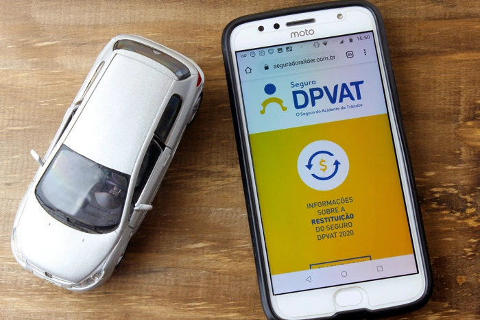 Miniatura de carro na cor prata ao lado de um celular com a logomarca do DPVAT na tela. Falando sobre seguro dpvat valor da indenização.