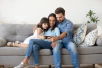 Família feliz acompanha algo em um smartphone enquanto está sentada em um sofá. Falando sobre seguro de vida de 500 mil.