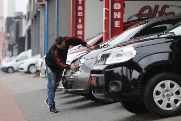 Carros estacionados em uma concessionária. Há uma pessoa limpando os faróis de um deles. Falando sobre seguradora para carros antigos.