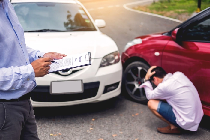 Profissional faz checagem de acidente de carro. Em segundo plano, dois carros colidem e homem lamenta agachado e com as mãos na cabeça. Falando sobre como funciona o seguro de carros para terceiros.